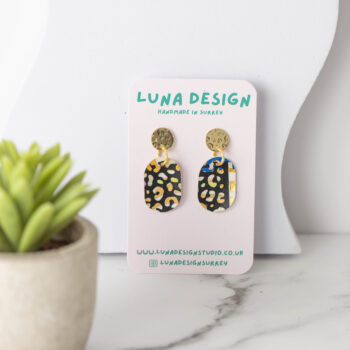 A pair of handmade earrings with gold circular studs and black oval drops featuring a yellow and white abstract pattern, displayed on a "Luna Design" card. The earrings are set on a white marble surface next to a small potted succulent.