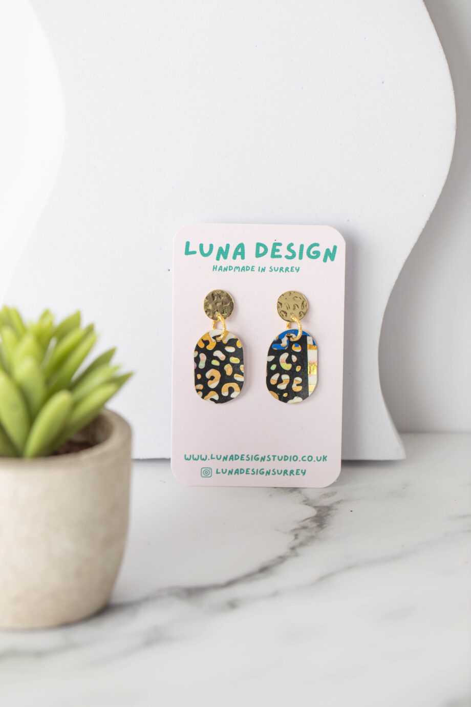 A pair of handmade earrings with gold circular studs and black oval drops featuring a yellow and white abstract pattern, displayed on a "Luna Design" card. The earrings are set on a white marble surface next to a small potted succulent.