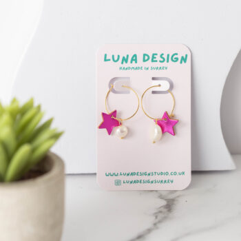 A pair of handmade earrings with gold hoops, each featuring a bright pink star charm and a small white bead, displayed on a "Luna Design" card. The earrings are set on a white marble surface next to a small potted succulent.