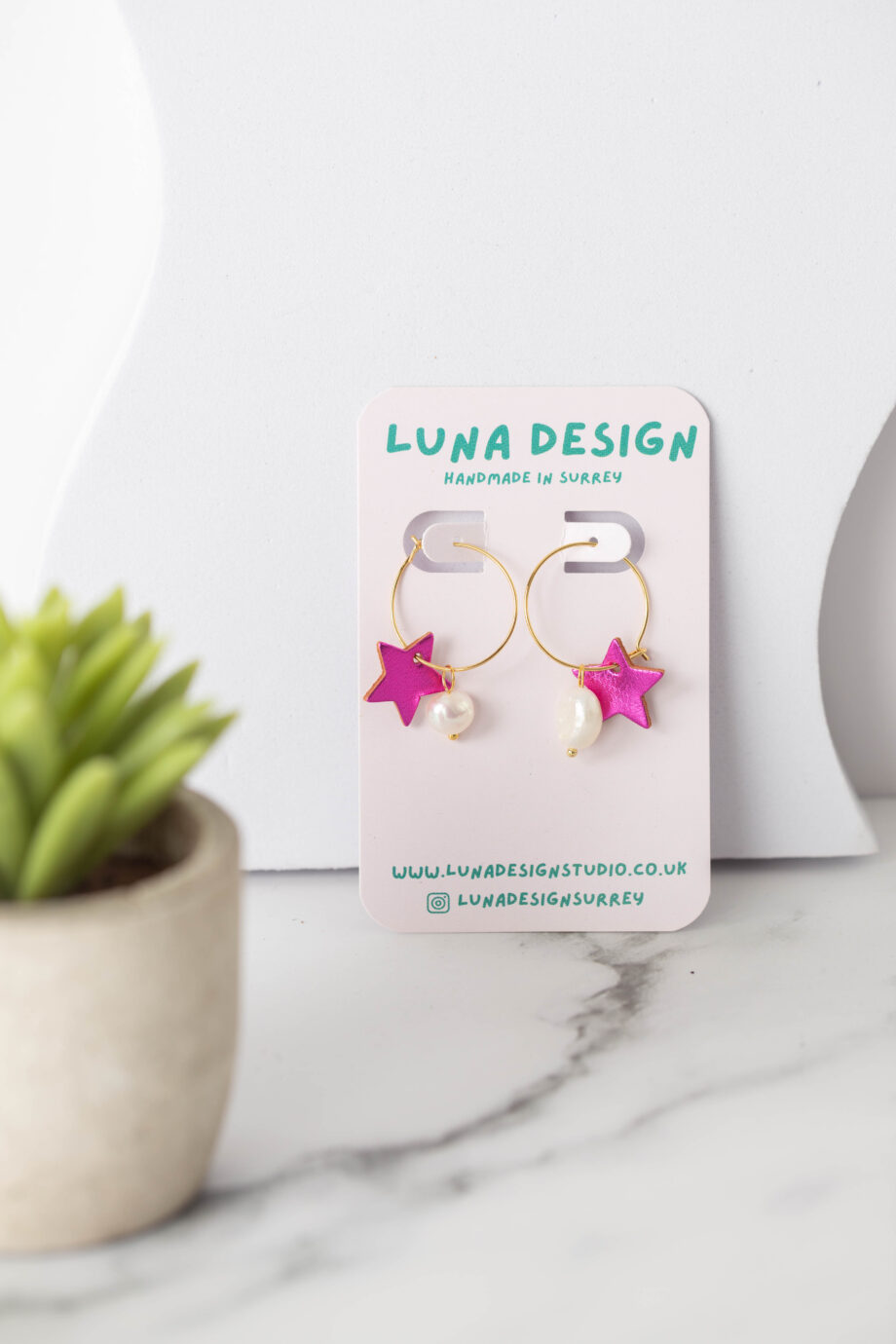 A pair of handmade earrings with gold hoops, each featuring a bright pink star charm and a small white bead, displayed on a "Luna Design" card. The earrings are set on a white marble surface next to a small potted succulent.