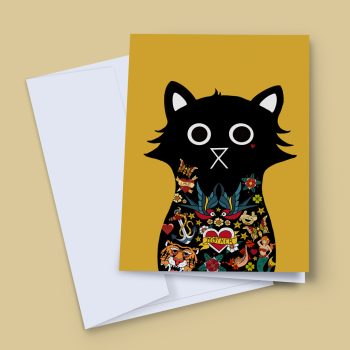 A 5x7 greeting card propped against a white envelope on a sandy beige background. The card features a bold black cat silhouette on a mustard yellow background, its body filled with colourful American traditional tattoo illustrations including swallows, an anchor, a tiger, a mermaid, a heart reading "Mother", flowers, and gold stars.