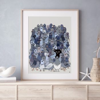 Framed illustrated art print of a large flock of grey and blue pigeons with a single black cat hidden among them, displayed in a light wood frame on a pale wooden sideboard with neutral home décor accessories.
